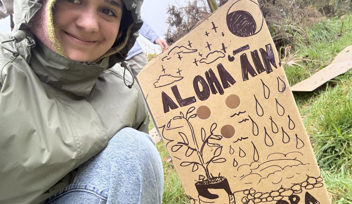 A selfie of Pono with a cardboard plant protector that has a drawing and the words "Aloha ʻĀina" and "Mauri Ora".