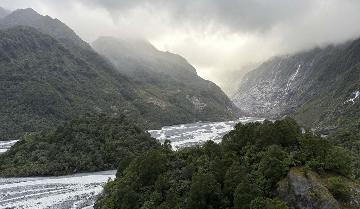 A massive landscape of mountains and valleys with small rivers running through and the sun peaking through the clouds.