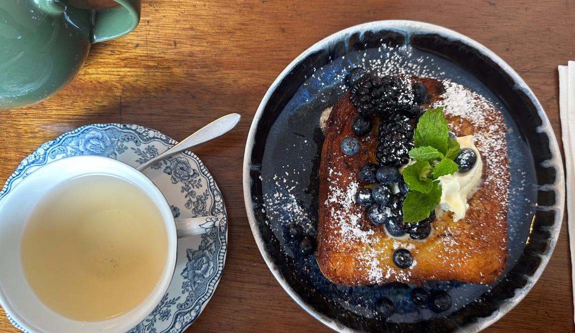 Image shows a cup of tea and a piece of french toast on a plate. The two items are next to each other.