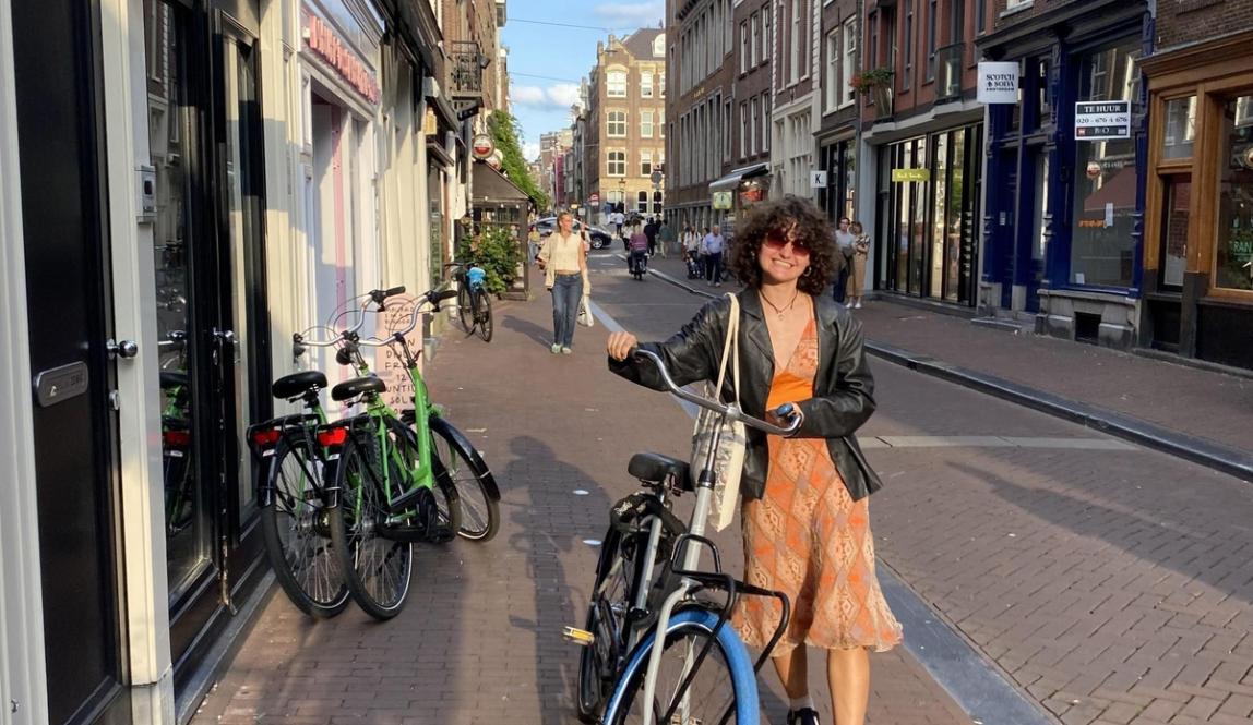 Student walking a bicycle along a brick-lined street in Amsterdam, with parked bikes, storefronts, and historic buildings stretching into the distance.