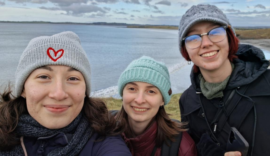 Three students standing on a coastal overlook in Ireland, with the shoreline and open water stretching into the distance behind them.