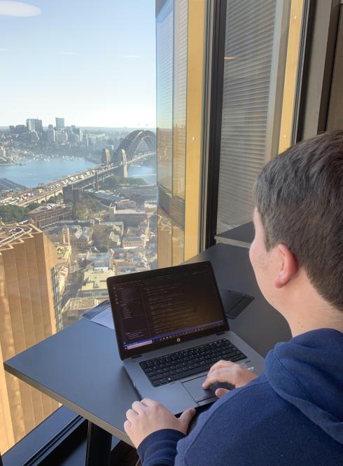 a student intern works on their computer while looking out the window at the Sydney Bridge