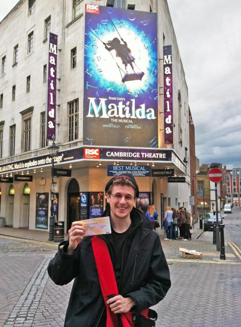 a male student standing outside of Cambridge Theatre with tickets to see the Matilda musical