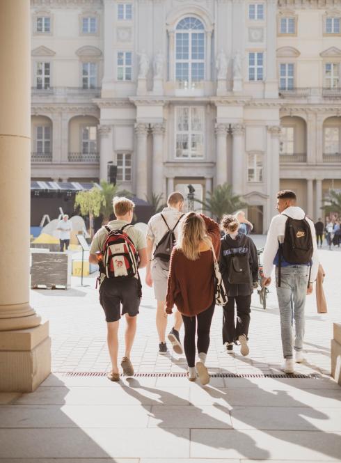 Students with their back toward the camera walk toward Humboldt Forum, a large white building, in Berlin.