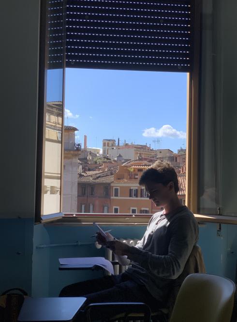 A student at the Rome Center works on their assignment with the window open. Outside the window are terracotta-colored buildings.