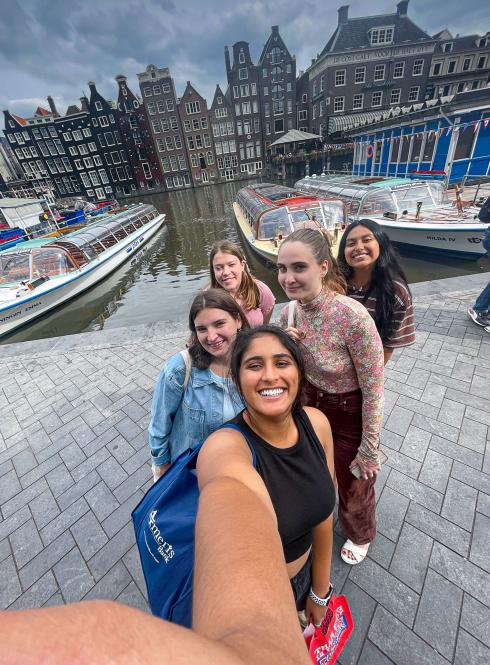five students standing in a line on cobblestone in front of a boat-filled amsterdam canal 