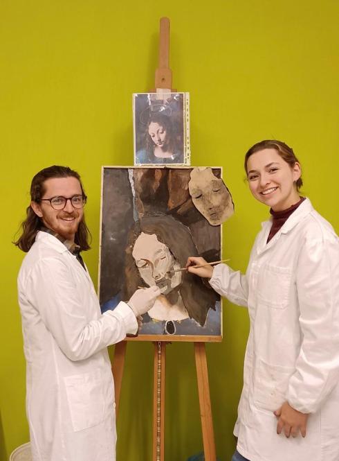 two students in white coats holding paint brushes in front of a painting on an easel