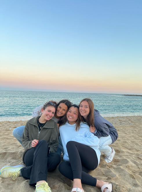 four students sitting on the sand at the beach with the ocean and the sunset in the background