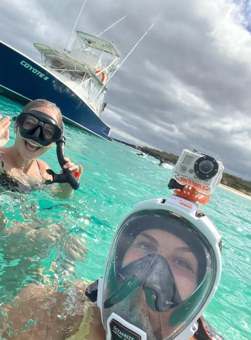 two students post for a photo in the ocean wearing snorkeling gear with a boat in the background