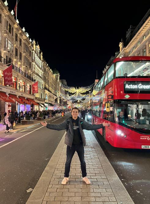 london student standing in street with arms stretched out next to red bus and string lighted buildings