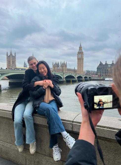 two students sitting on ledge hugging while another student holds up a camera and takes a picture