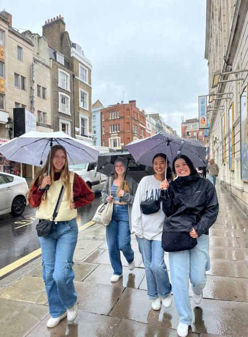 four students holding umbrellas walking down a sidewalk with tall windowed buildings in the background