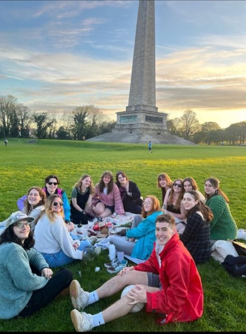 group of students sitting on the grass with picnic stuff in front of a stone monument