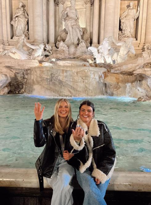 Two students sitting on the edge of the Trevi Fountain at night laughing, throwing coins behind them.