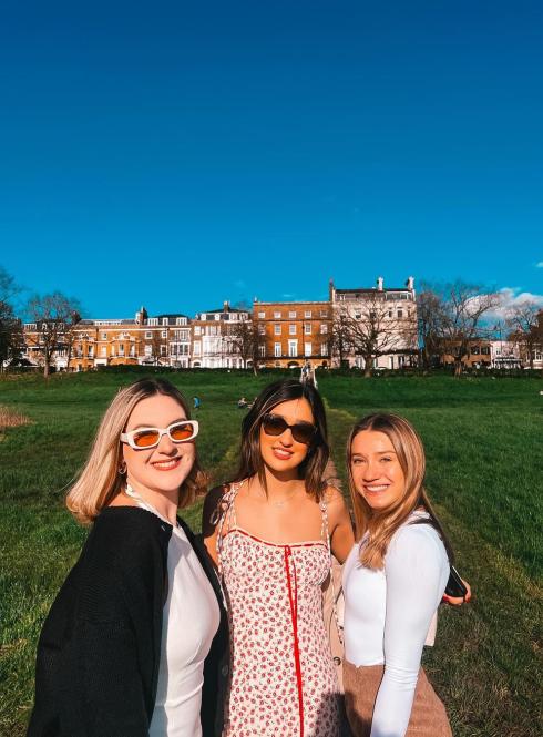three students standing on grass in sunlight in front of buildings