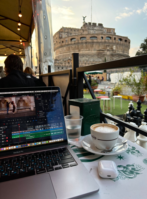 A cup of coffee and a laptop with video editing software at a cafe in front of the Colosseum.