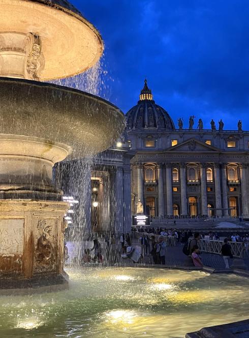 A nighttime photo of a fountain lit up in front of the Vatican.