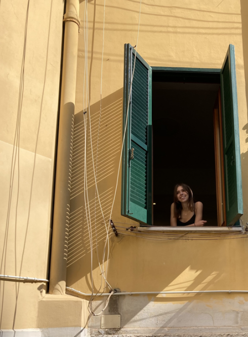 A woman looks out of a window with green shutters against a yellow building.