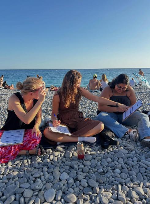 three students sitting on beach with clip boards