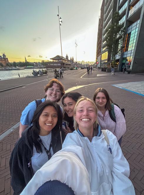 Five students taking a selfie along Amsterdam’s waterfront at sunset, with the water, historic buildings, and city skyline visible in the background.
