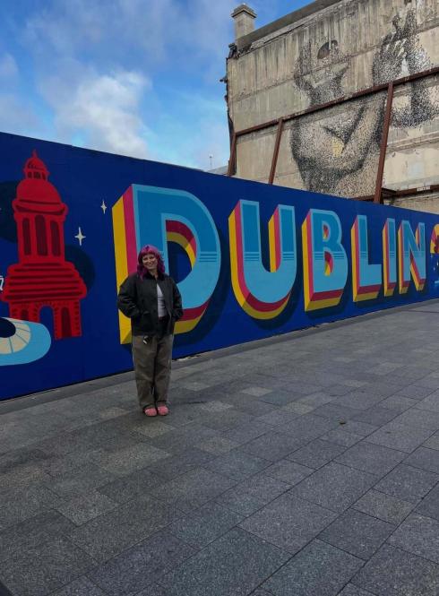Student posing beside a large “Dublin” mural in the city center, with a historic building mural painted on a nearby wall.