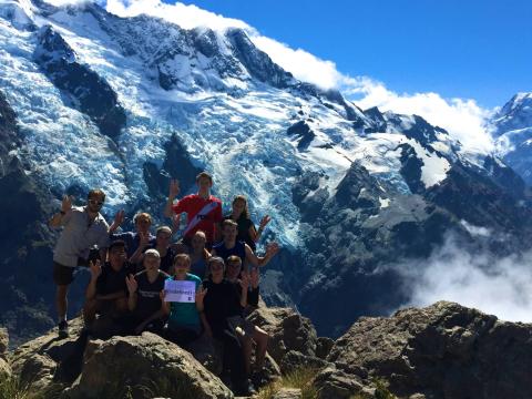 Group of hikers posing on a rocky mountain, snowy peaks in the background under clear blue sky. This alt text was added with Al; accuracy may vary.