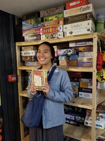 Brittany in front of a bookshelf filled with games. In her hand she holds a box of the game named "Bang!"