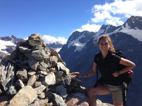 A person with a backpack stands smiling on a rocky mountaintop with snowy peaks in the background. This alt text was added with Al; accuracy may vary.