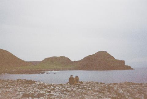 A couple sits looking at one another across from a landmass on the Giant's Causeway. The image is grainy.