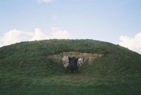 An image of a green burial mound with an opening, in front of which a person is kneeling to look in