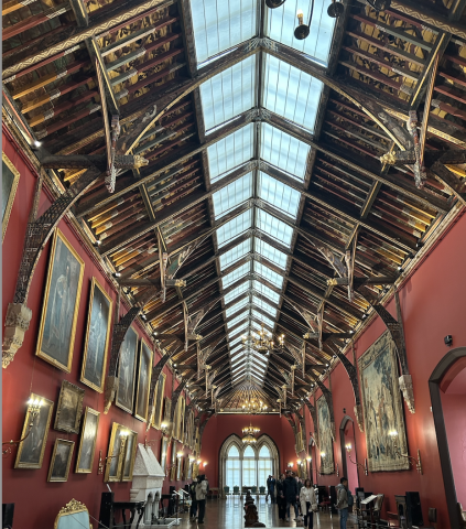 A long gallery with red walls and paintings hanging. The arched ceiling is made up of wooden beams painted with patterns, animals, and figures.