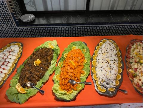 Buffet-style spread of traditional Moroccan foods on silver plates, including zaalouk, marinated carrot slices, and white rice with raisins. Each dish is decorated with a frame of thin orange slices.