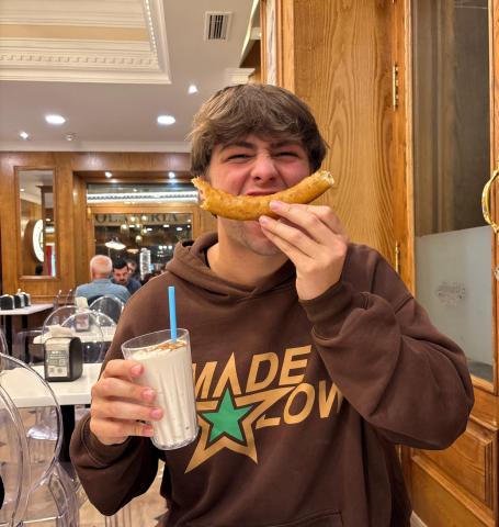 My friend Caden sits inside the churro shop at night, smiling. His face is scrunched up and he holds a curved churro in front of his mouth to mimic a smile, clutching a cinnamon-dusted leche rizada in the other hand.