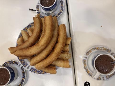 10 churros stacked on a plate in the center of the table, surrounded by three steaming cups of thick hot chocolate in ceramic teacups.