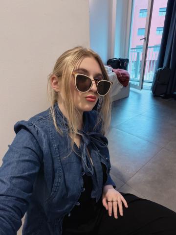 The author of the blog wearing a denim long sleeve shirt and black pants. She is siting on the floor and the camera is angled to show the open balcony door of the dorm room she is in.