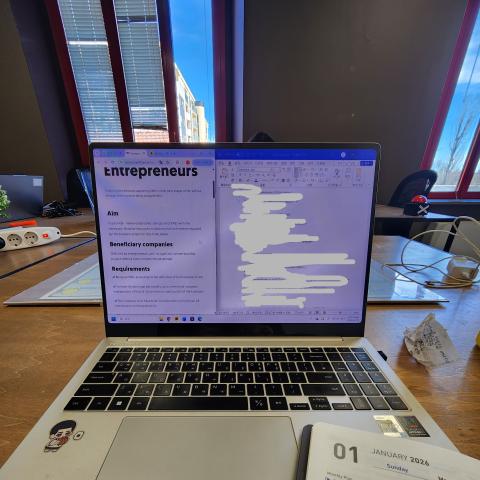 A laptop showing research documents on a desk, with a 2026 planner open beside it in an office setting.
