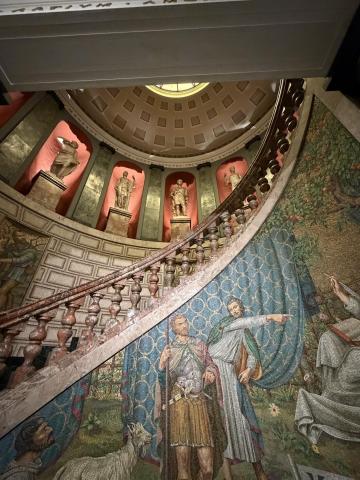A curved staircase with a mosaic embedded in the banister. It shows multiple men discussing something in front of a blue curtain.