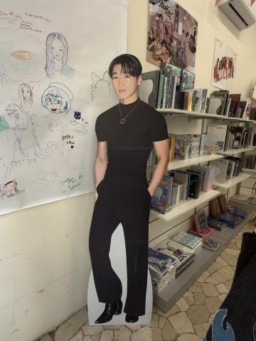 A photo of a cardboard cutout of San from Ateez. He is wearing a black shirt with black pants. In the background is a shelf full of colorful albums. 