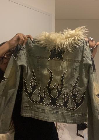 Me holding up a jean jacket with tan faux fur lining the collar and and black and silver detailing creating flames on the back.