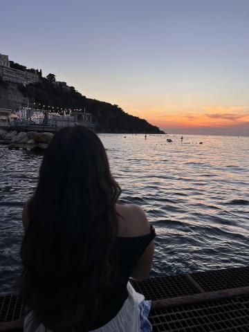 A person sits by the water in Sorrento, watching the sunset over the sea as the sky is pink and orange. 
