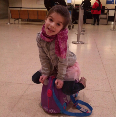 Young girl smiling while sitting on a small purple ride-on suitcase in an airport terminal.