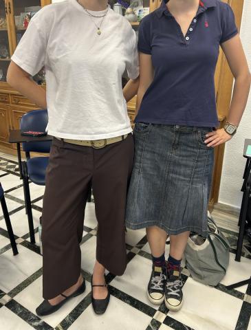 Two students. The student on the left is wearing brown pants, flats, and a white tee shirt. The student on the left is wearing a blue short sleeve polo, a calf length denim skirt, and converse.