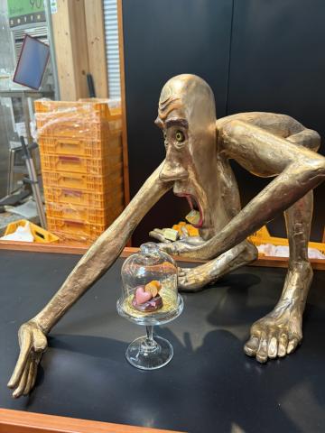 A metallic statue of a humanoid figure with long limbs crouches over a chocolate as if about to eat it.