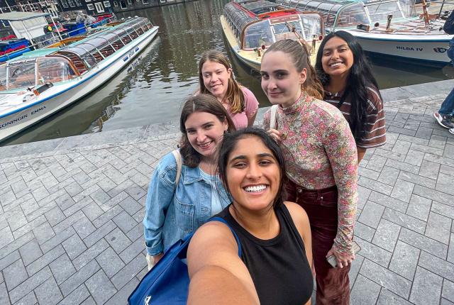 five students standing in a line on cobblestone in front of a boat-filled amsterdam canal 