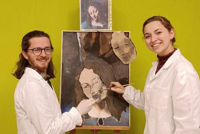 two students in white coats holding paint brushes in front of a painting on an easel