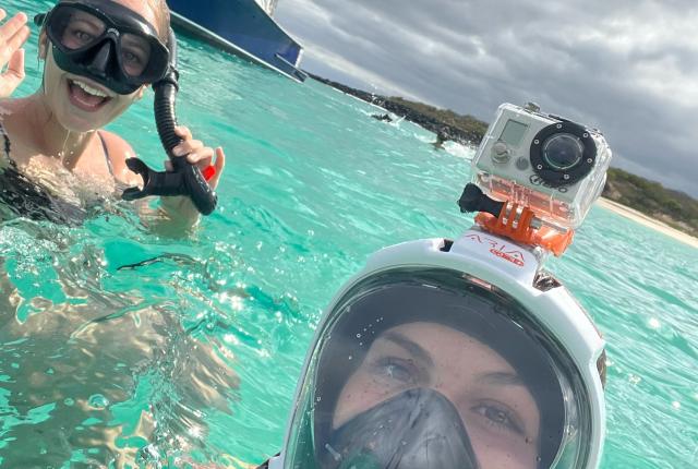 two students post for a photo in the ocean wearing snorkeling gear with a boat in the background