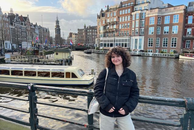 Student posing on a bridge above an Amsterdam canal, with a tour boat, waterfront buildings, and city landmarks visible behind them.