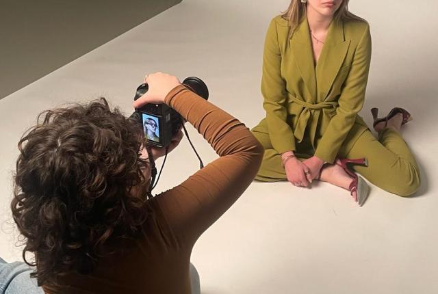 A fashion photoshoot with a model in a green pantsuit sitting on the floor, with the photographer sitting in front of them taking their photo