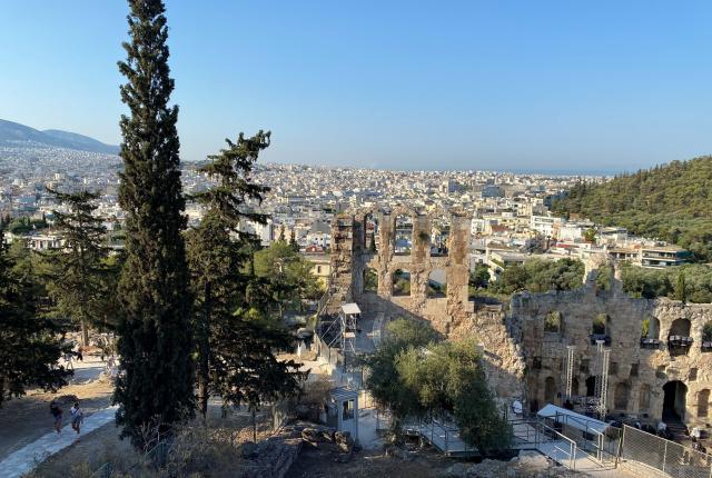 View of Acropolis ruins with cypress trees and cityscape in the background. This alt text was added with Al; accuracy may vary.