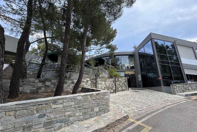 Modern building with stone walls, glass facade, surrounded by trees under a cloudy sky. This alt text was added with Al; accuracy may vary.
